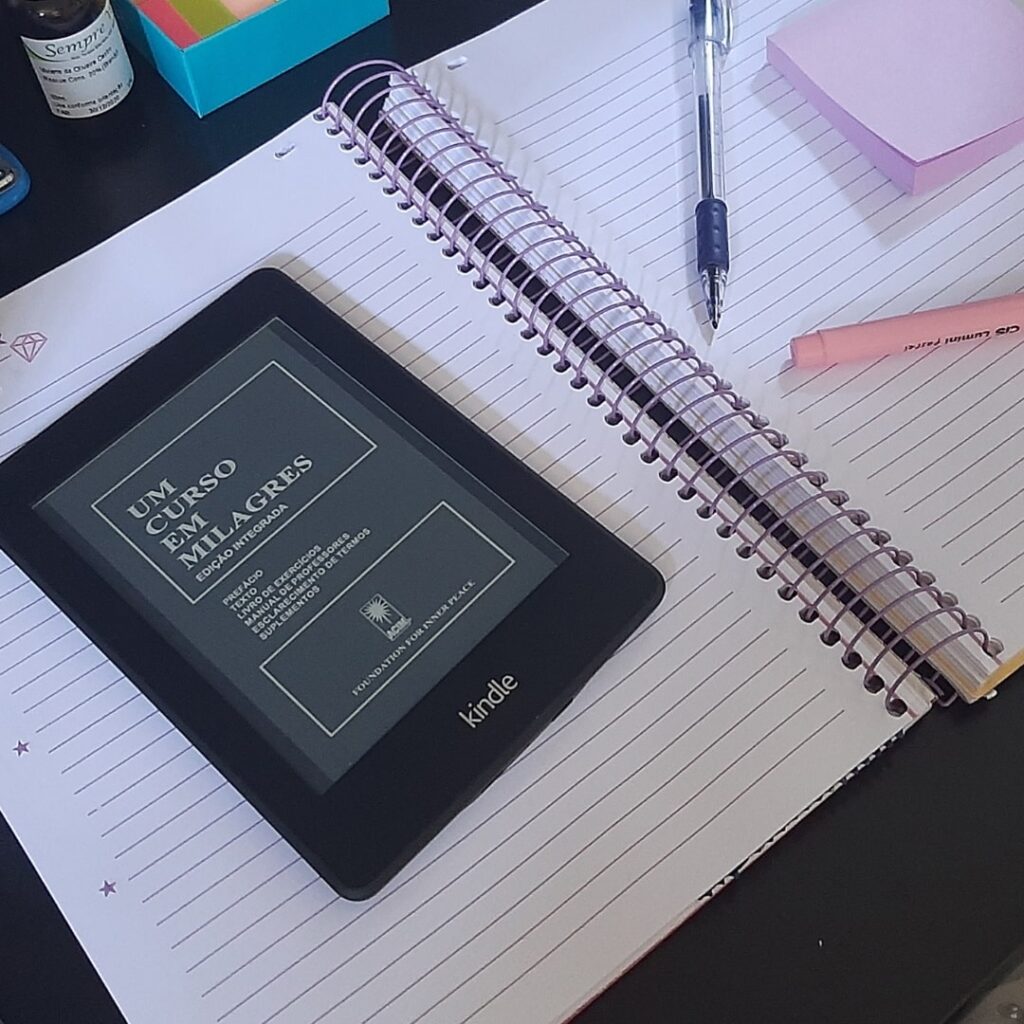 kindle com o livro um curso em milagres em cima de um caderno com canetas e post-its